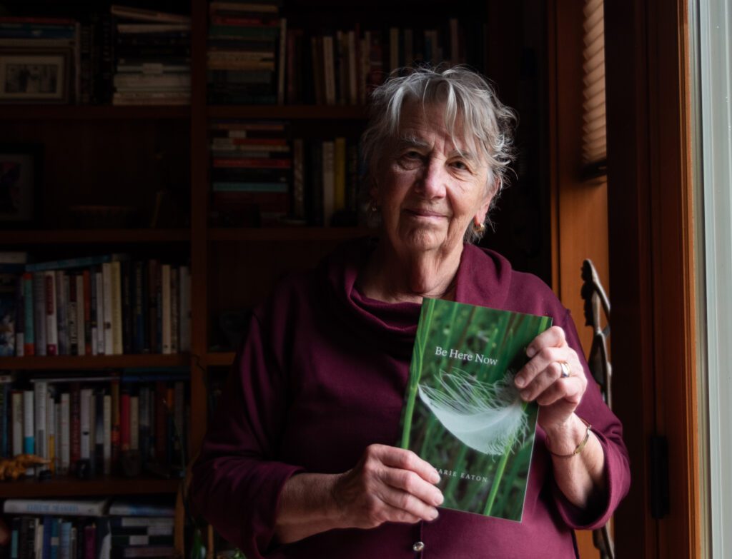 Marie Eaton holds a copy of her upcoming collection of poems "Be Here Now" where the book cover is a feather falling in green foliage.