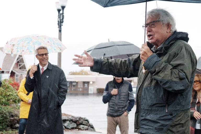 Brian Griffin, right, speaks to a crowd during the unveiling of a street sign near the Fairhaven Library as all attendees brave the rain by using umbrellas.