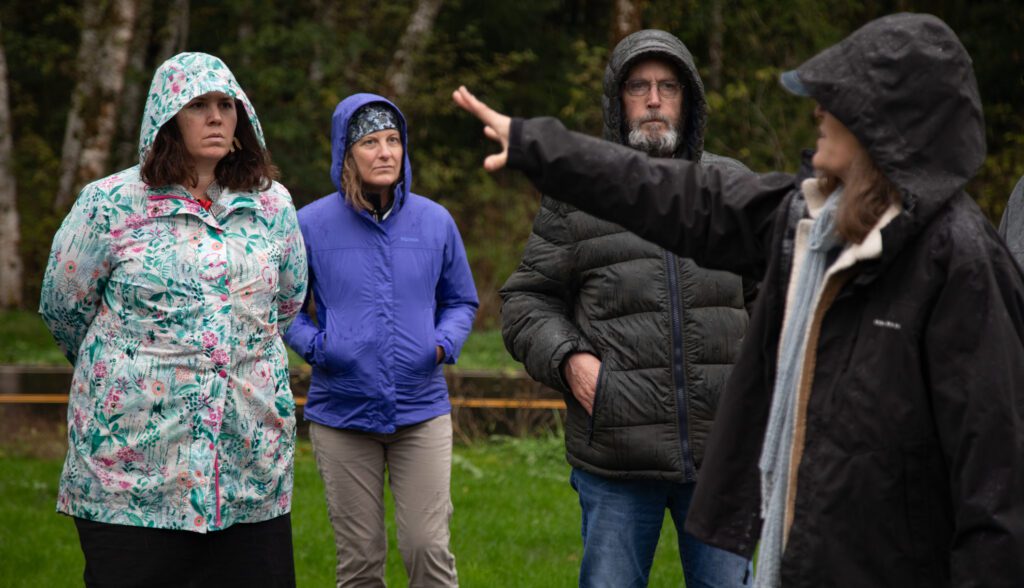 From left, state Sen. Sharon Shewmake and Whatcom County Council members Carol Frazey and Barry Buchanan listen to Paula Harris talk as she gestures to the left while wearing wet rainproof gear.