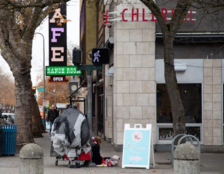 A person sits on the corner of Holly Street and Railroad Avenue next to a colorful sign as they sit next to a shopping cart full of items.