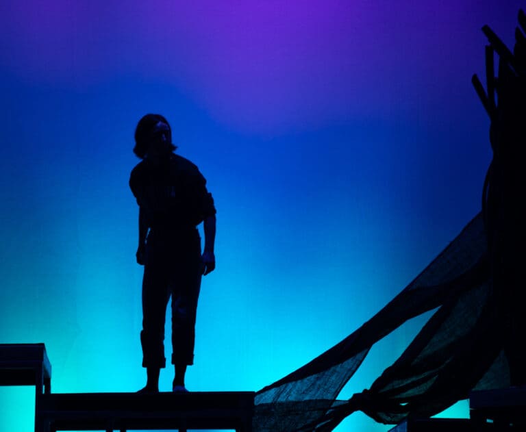 Jackson van Pelt performs as Puck on stage with a blue-purple gradient behind them.