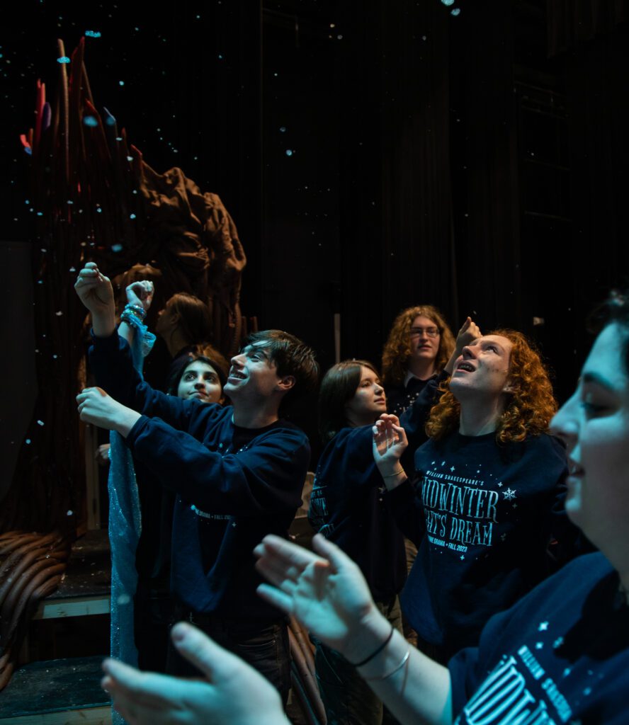 Actors and stage crew dance and laugh underneath the "snow" (soap bubbles) while dressed in the show's title and themed sweater.