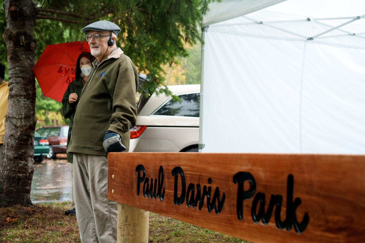 Paul Davis, 97, stands next to the sign for the newly named Paul Davis Park with headphones on next to a woman with a bright red umbrella.
