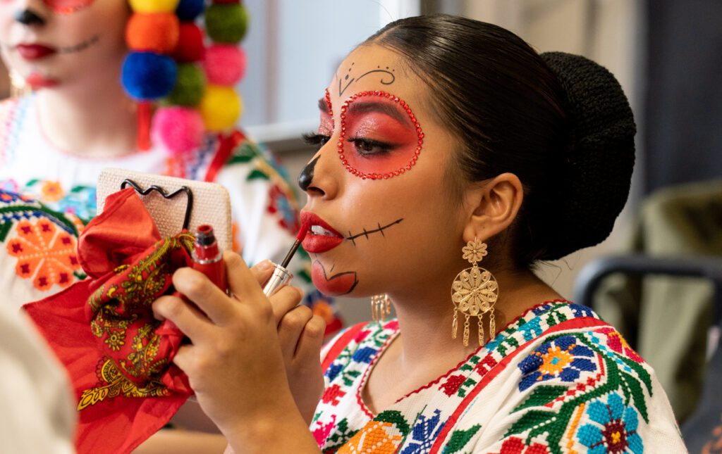 Arianna Gonzalez applies lipstick while using a small mirror next to another dancer dressed in a similar colorful attire.