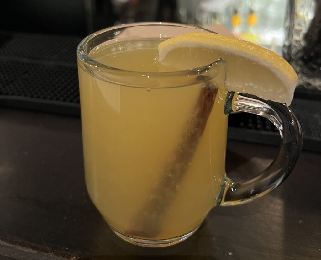 The Naughty Toddy at Penny Farthing is a yellow drink served in a mug and garnished with a stick of cinnamon and a slice of lemon.