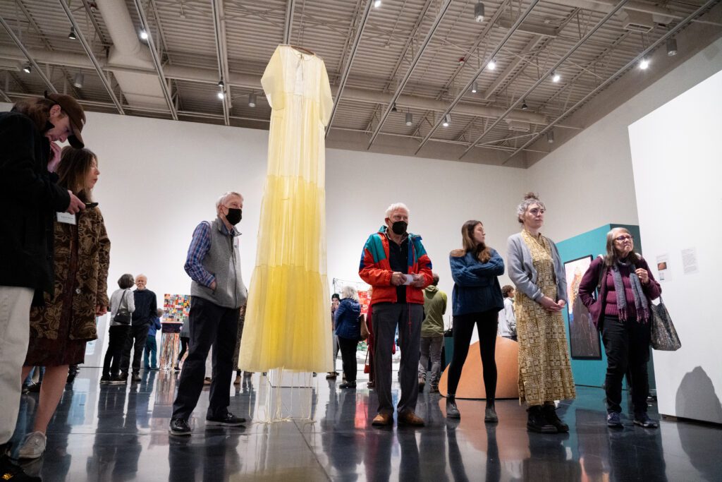Margaret Chodos-Irvine's piece : "PPE: 14 Days With My Father in Assisted Care in 2020," is a long beige gown that hangs from the ceiling anext to attendees who are looking at another art piece.