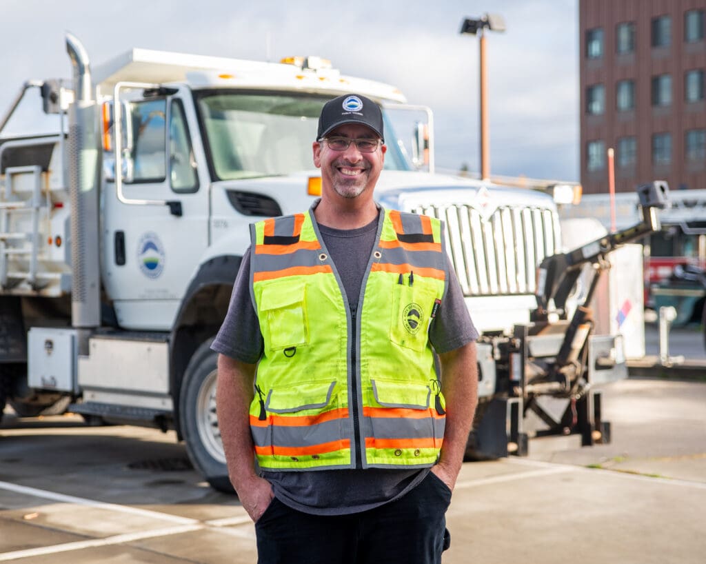 Scott Van Diest stands in front of a City of Bellingham dump truck wearing a safety vest with his hands tucked in his pockets.