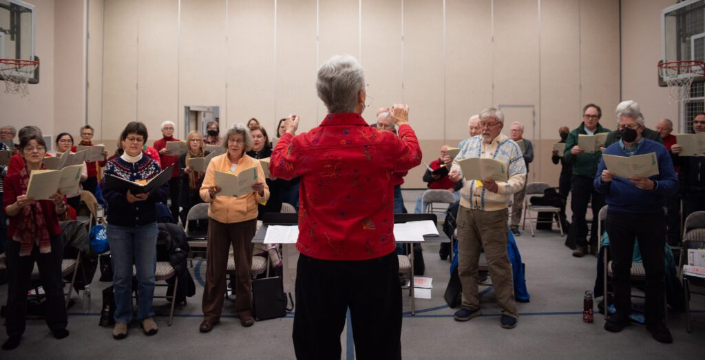 Deborah Brown conducts the Whatcom Chorale as they gesture behind a podium.