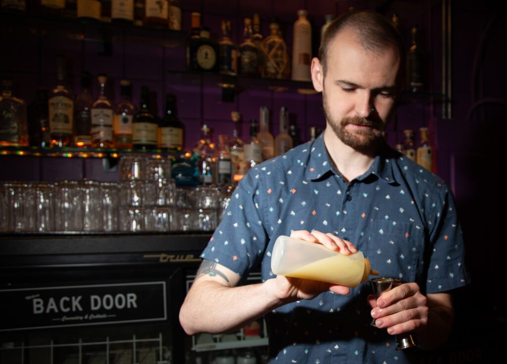 Erik Tostenrude, in front of a shelf full of drinks and glass, squeezes a shot of passionfruit-curry puree into a measurer for the drink Blackjack.