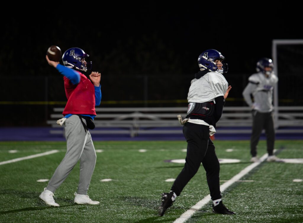 Quarterbacks freshman Ryan Harrington, left, and senior Rex Larson throw passes during practice as one player is dressed in red and blue.