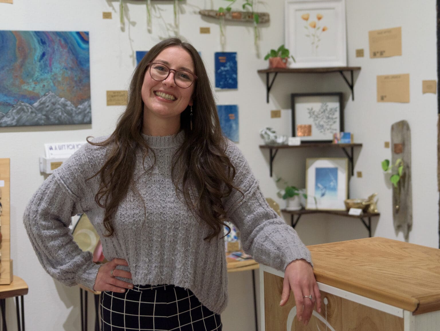 Paige Woods stands in the Heron's Nest art gallery leaning on the wooden counter as she smiles for the camera.