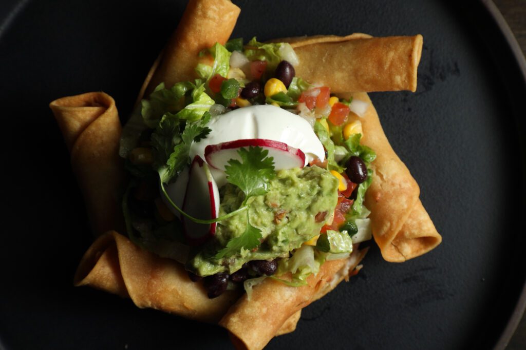 Rolled chicken tacos surround a bed of southwestern salad topped with sour cream and guacamole are severed on a black plate.