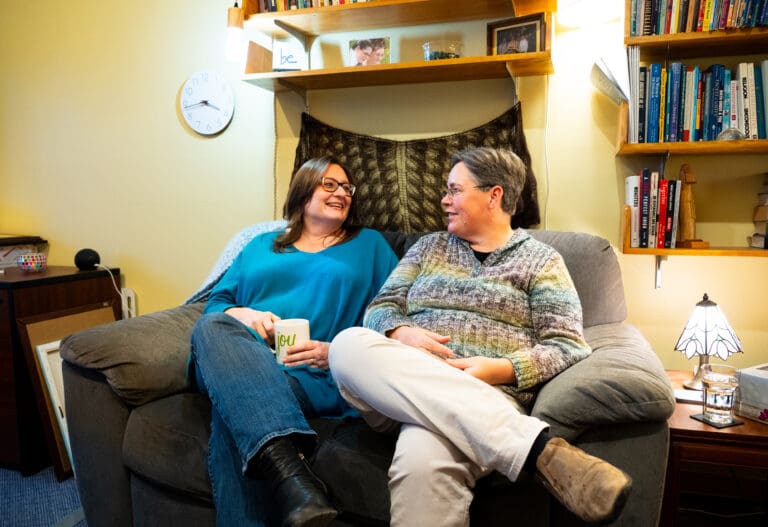 Two women wearing glasses sit on a couch with their legs crossed and smile at each other.