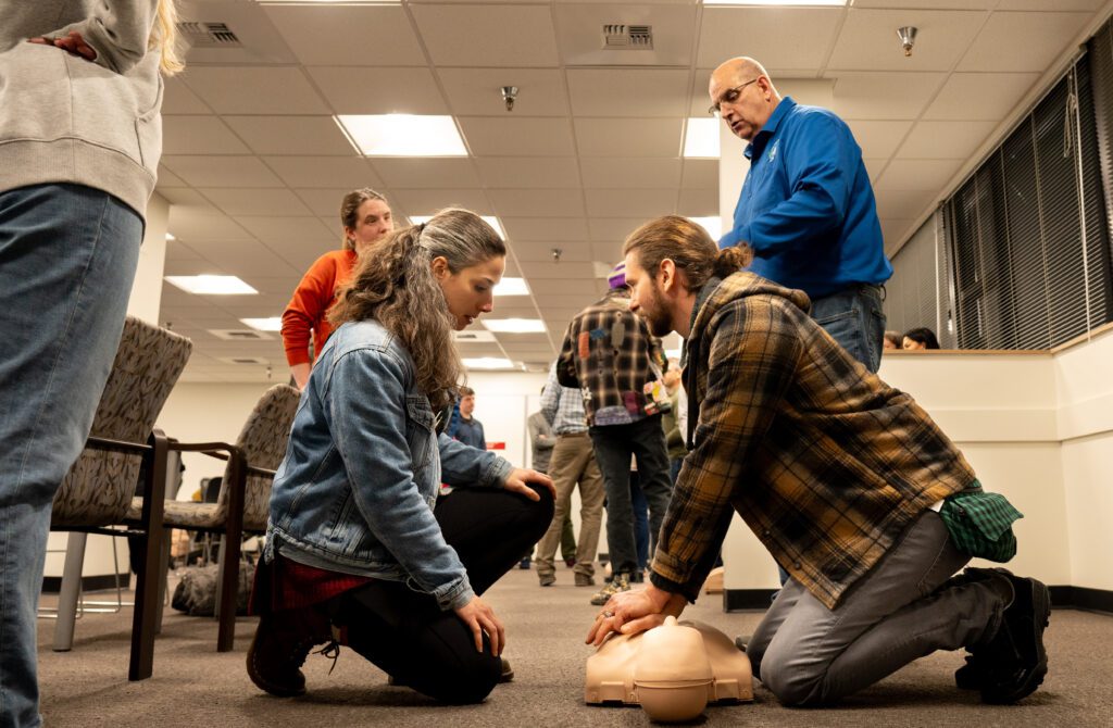 Shannon Bossi, left, and her husband, Steve Moore take turns performing chest compressions on a dummy as an instructor stays close to give instructions while others are seen in the background, chatting.