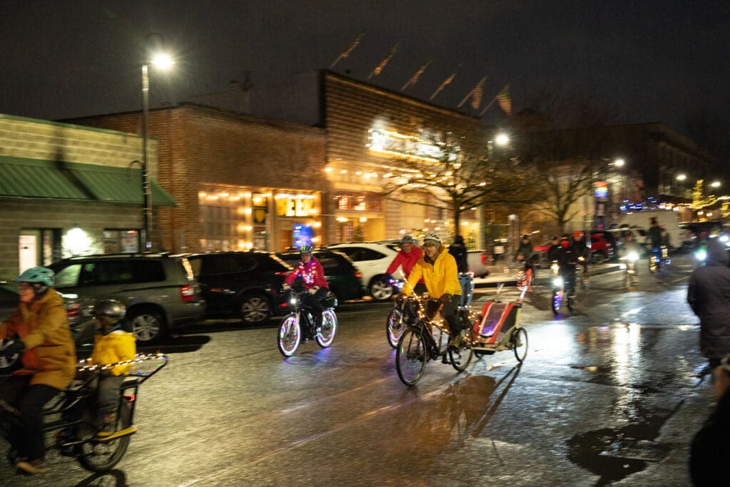 Cyclists on the main roads, all of which are wearing safety gear and various different rain gear and bright lights decorated on their bikes.