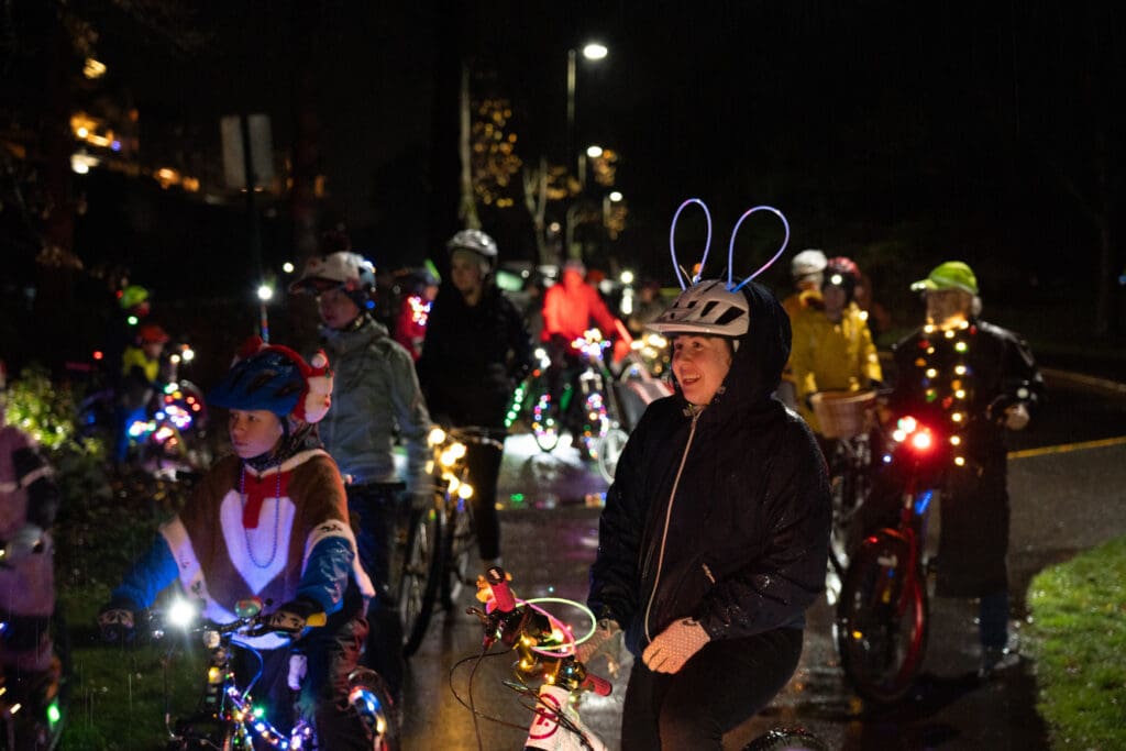 Cyclists wearing bright clothing and lights as well as their bicycles donning bright lights gather at Boulevard Park. A woman in the front has bunny ear lights on her helmet.