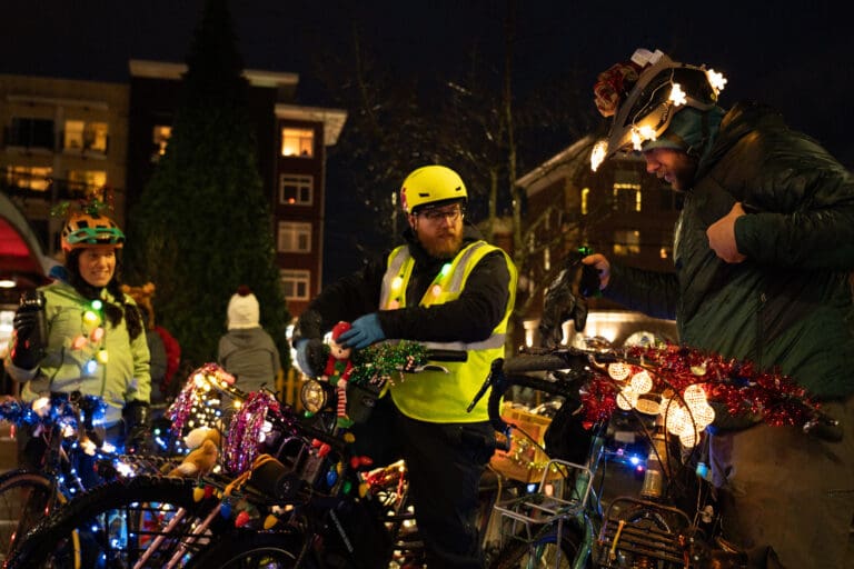 Deanna Ciaccia, left, Kevin Roemer, and Andrew Stuntz chat while wearing safety vests, waterproof clothes, and bicycle helmets decorated with bright lights as their bikes are decorated with the same multicolored decorations and lights.