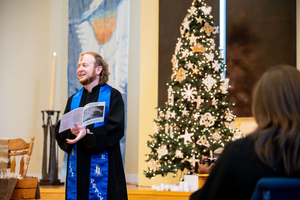 A man wearing a blue pastor stole stands in front of a Christmas tree holding a paper as he gives a sermon.
