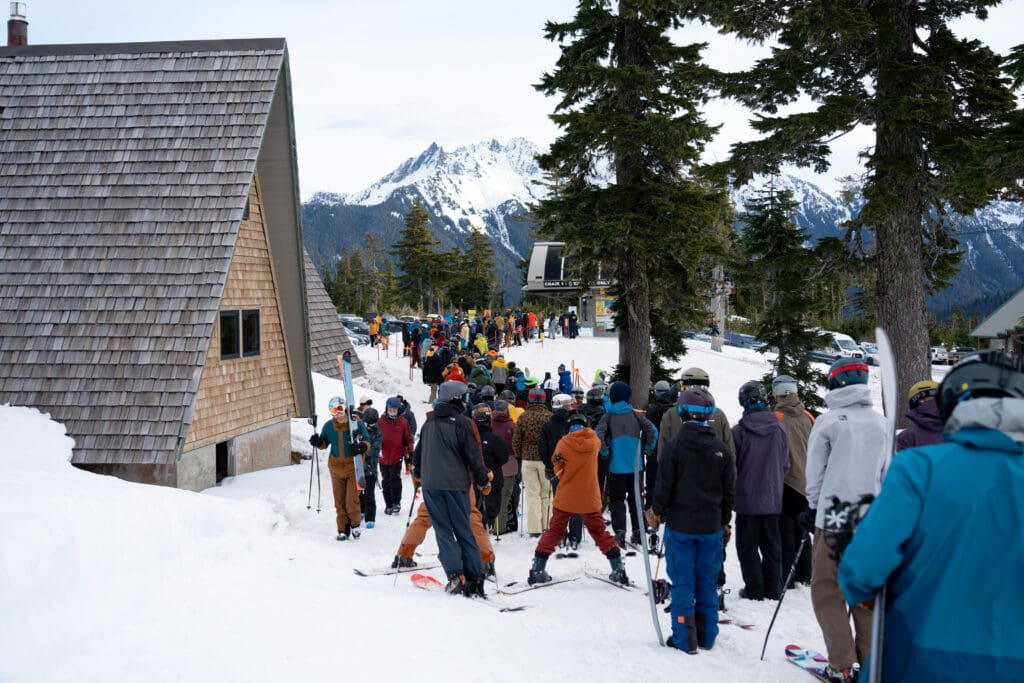 Skiers and boarders form a line at Chair 1 at Mt. Baker Ski Area, dressed in different snow attire and holding onto different snow gear.