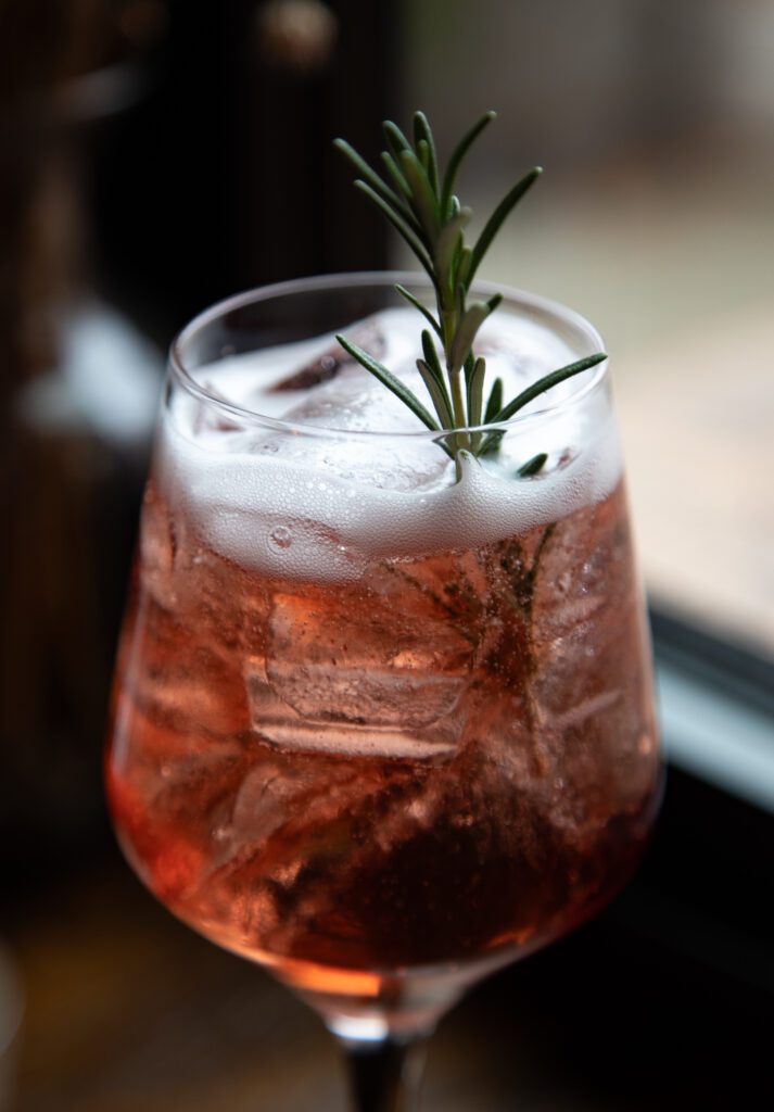 The Jack of Hearts cocktail is a rosey-red drink poured in a wine glass garnished with rosemary.