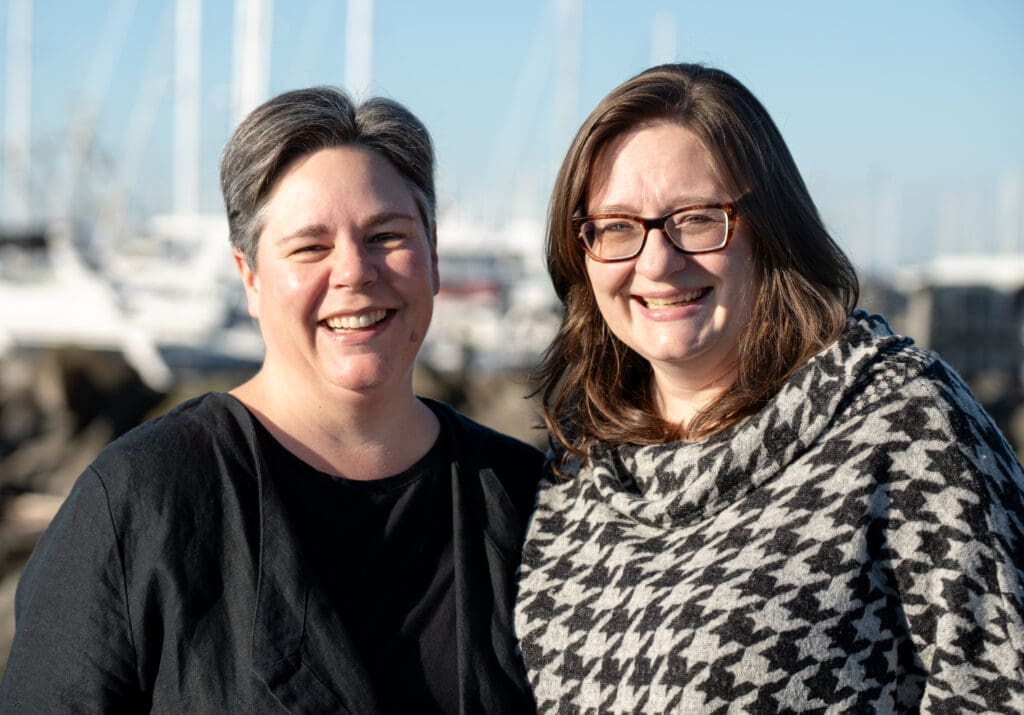 Two women smile and pose for the camera with sailboats in the background.