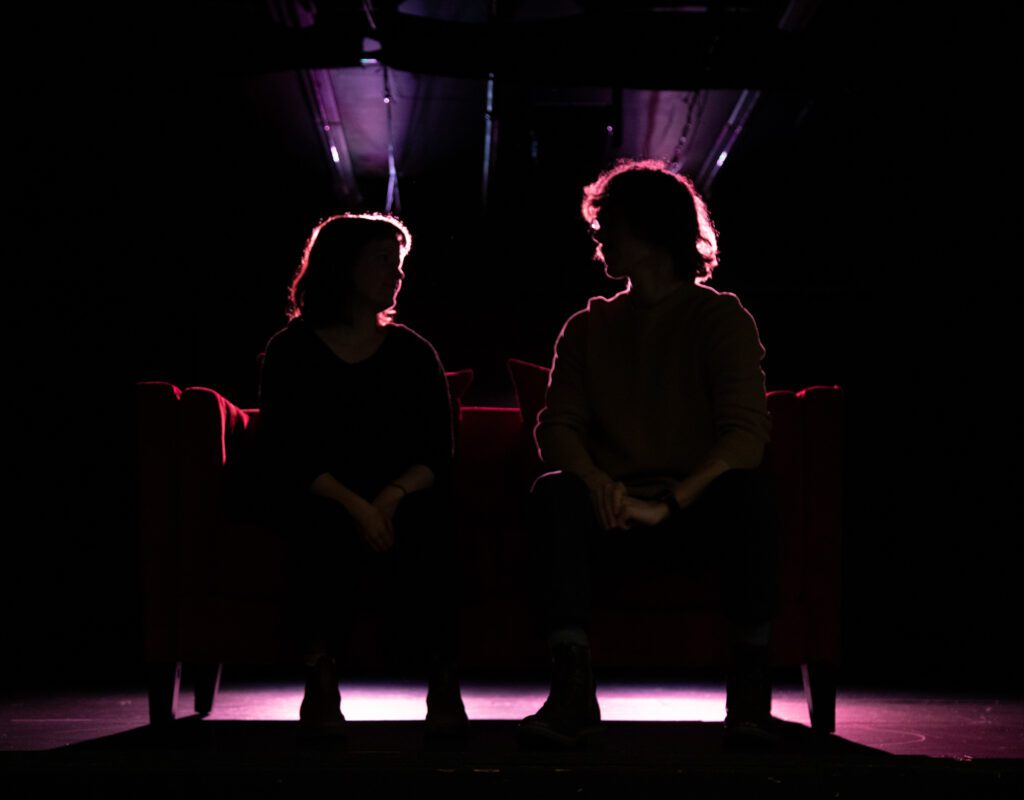 Juliette Machado, left, and Kaleb Van Rijswijck sitting next to each other on a sofa as a bright light shines on them from behind them.