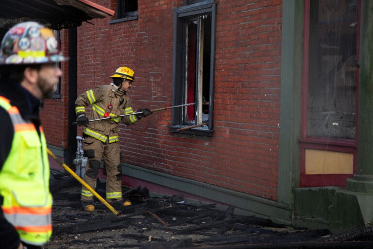 A firefighter breaks a window with a metal rod as another person walks by wearing a safety vest and helmet.
