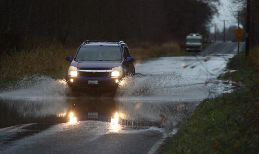 A car drives through standing water on Ferndale Road, splashing the sides with water from the drive.