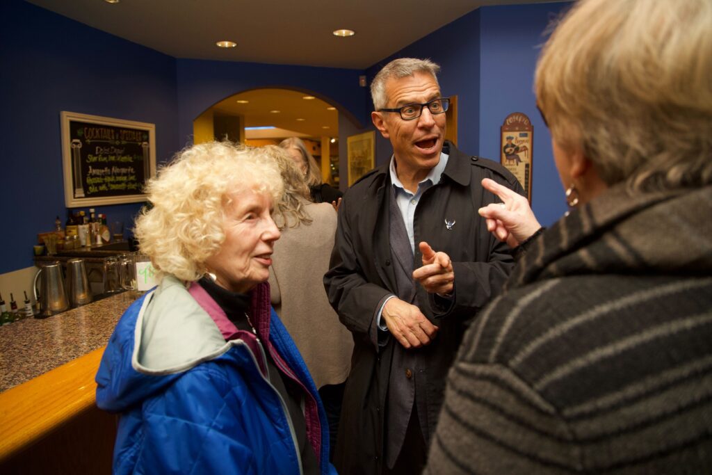 Former city council member Louise Bjornson, left, and Seth Fleetwood surrounded by people in a blue room as a woman points back at him.