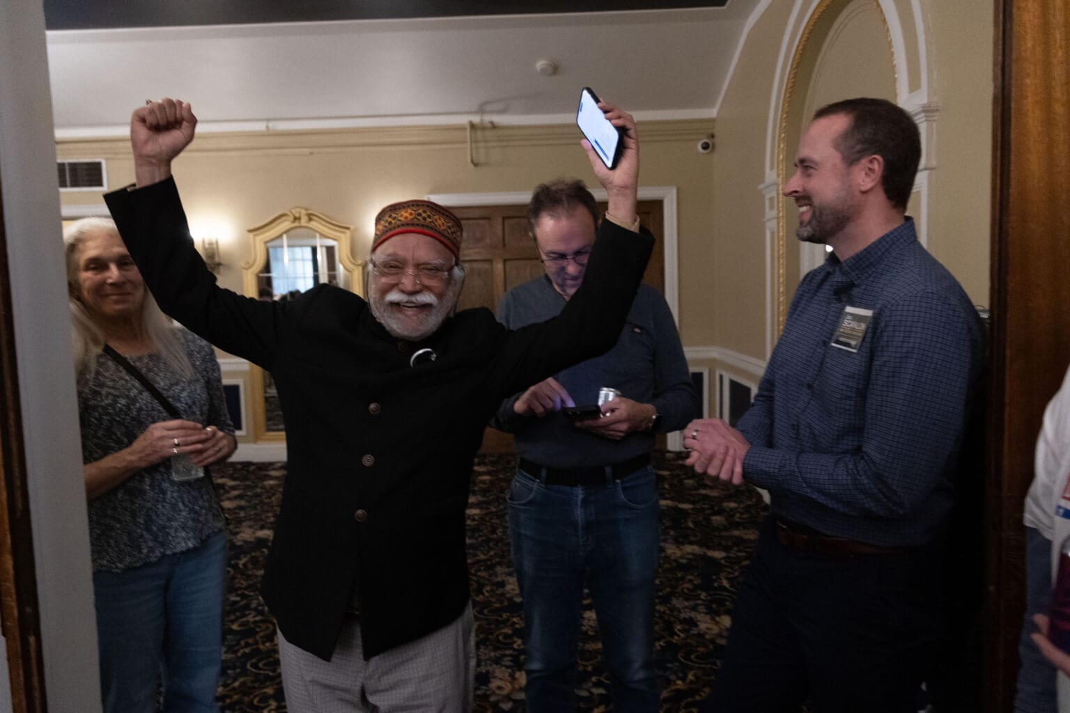 Whatcom County Executive incumbent Satpal Sidhu reacts in celebration with a phone in hand and both arms raised.