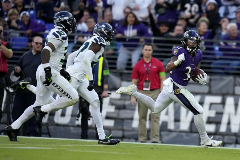 Baltimore Ravens running back Keaton Mitchell runs with the ball away from two defenders trailing right behind.
