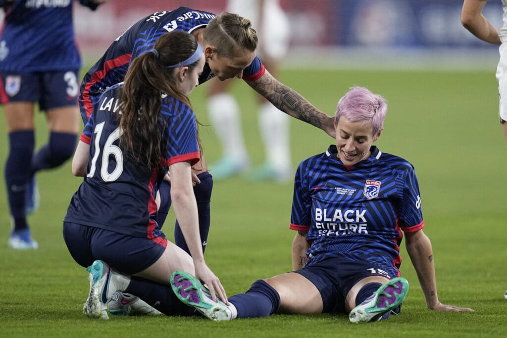 OL Reign forward Megan Rapinoe, right, stays seated on the grass as two teammates console her.