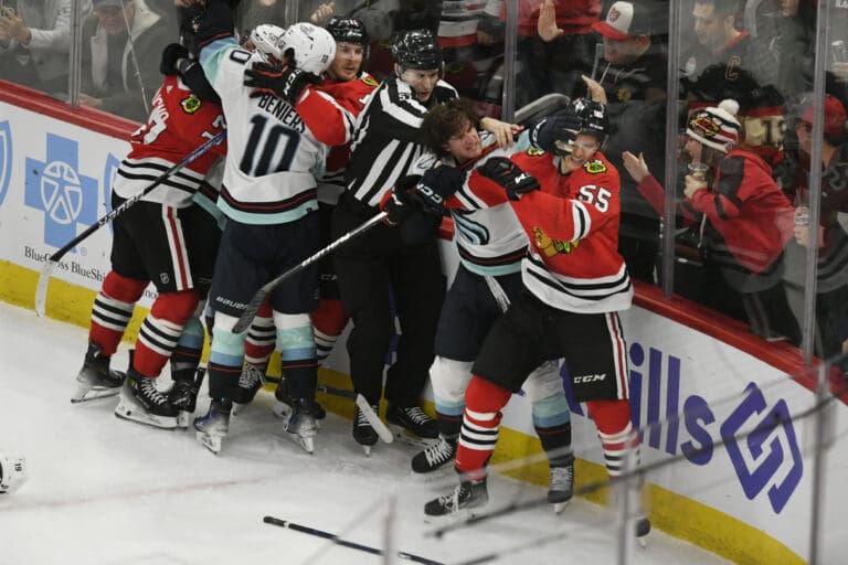 Players from the Chicago Blackhawks and Seattle Kraken fight against the glass barrier as spectators watch and take pictures of the struggle.