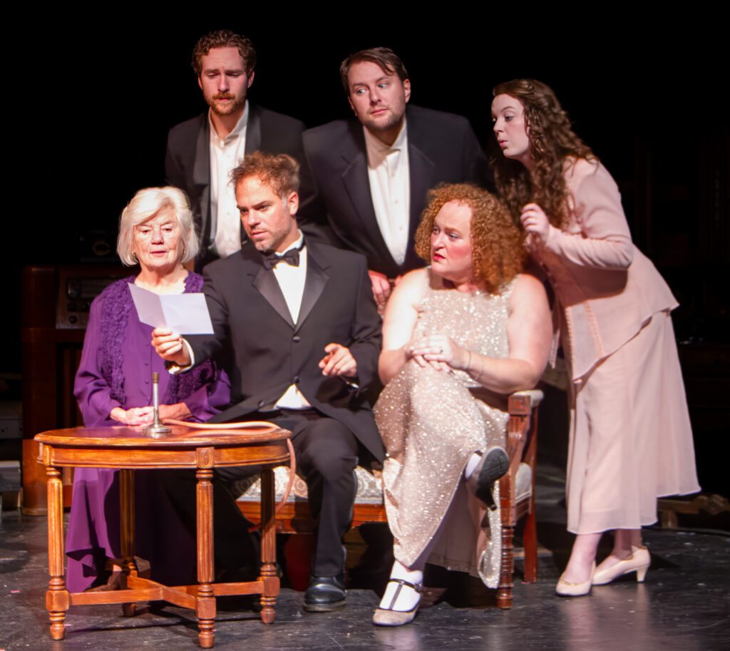 “The Game’s Afoot's" cast performing on stag where they are all looking at a piece of paper held up by a performer.