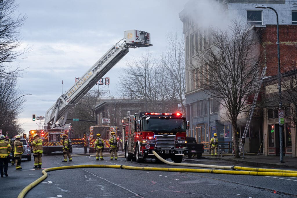 A crowd of firefighters discuss as a firetruck extends its ladder platform towards the smokey building.