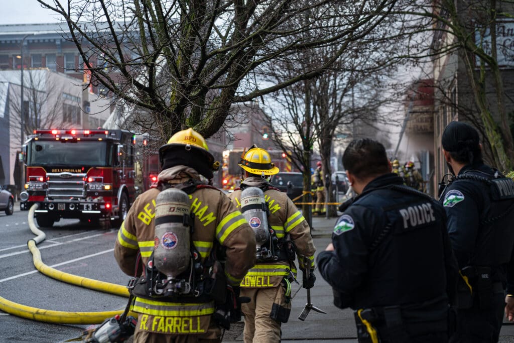 Bellingham Police officers look on as firefighters dressed in full gear pass by them on the sidewalk.