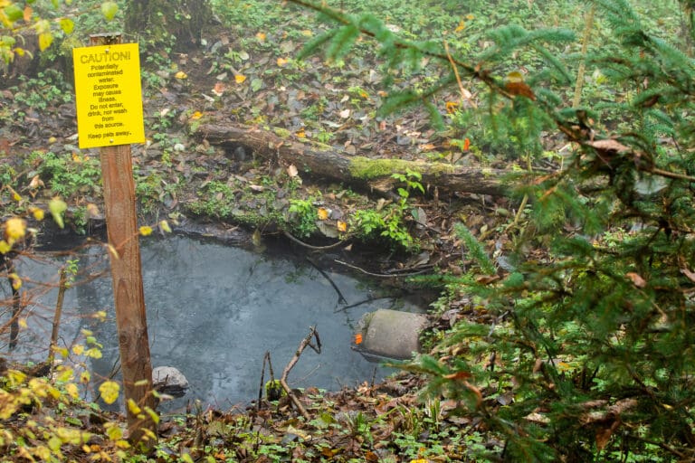 A bright yellow sign erected next to a small pool of water where a sewage pipe leaks into is warning to be cautious of the portentially contaminated water.