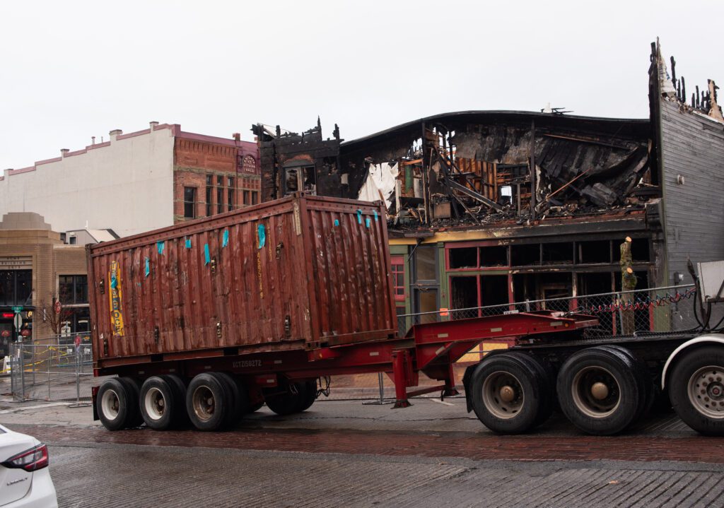 A maroon metal trailer is parked next to the burned down building.