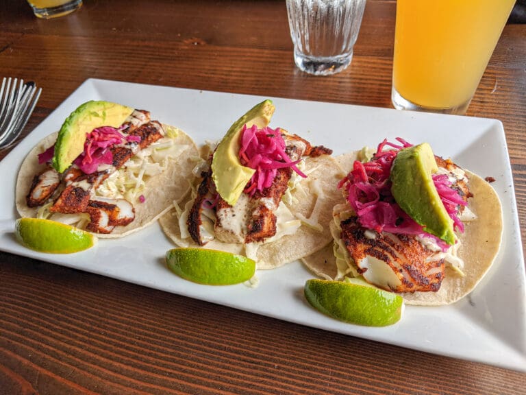 A row of fish tacos served with slaw, pickled vegetables and garnished with lime slices.