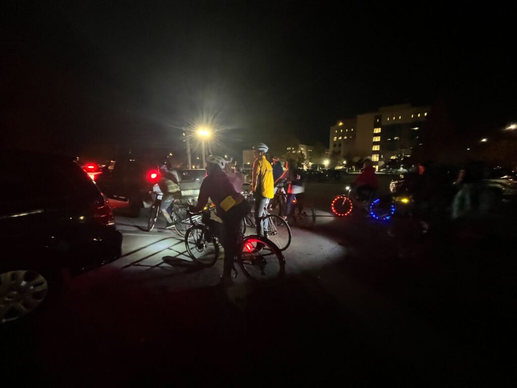 The Black Cat bicyclists ride by the ballot box wearing safety cycling equipment with one participant decorating their bike with neon lights.
