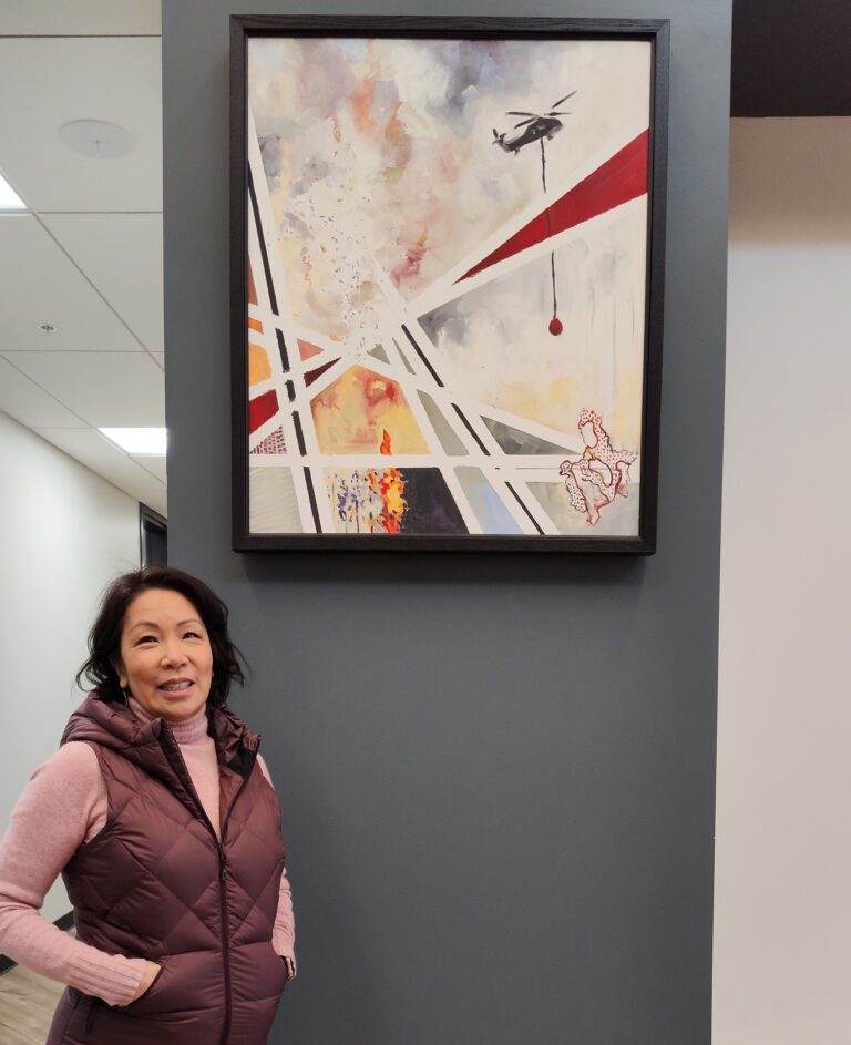 Soo Ing-Moody standing next to a painting by artist Kayla White, depicting the 2014 Carlton Complex wildfire with abstract shapes, colors and lines.