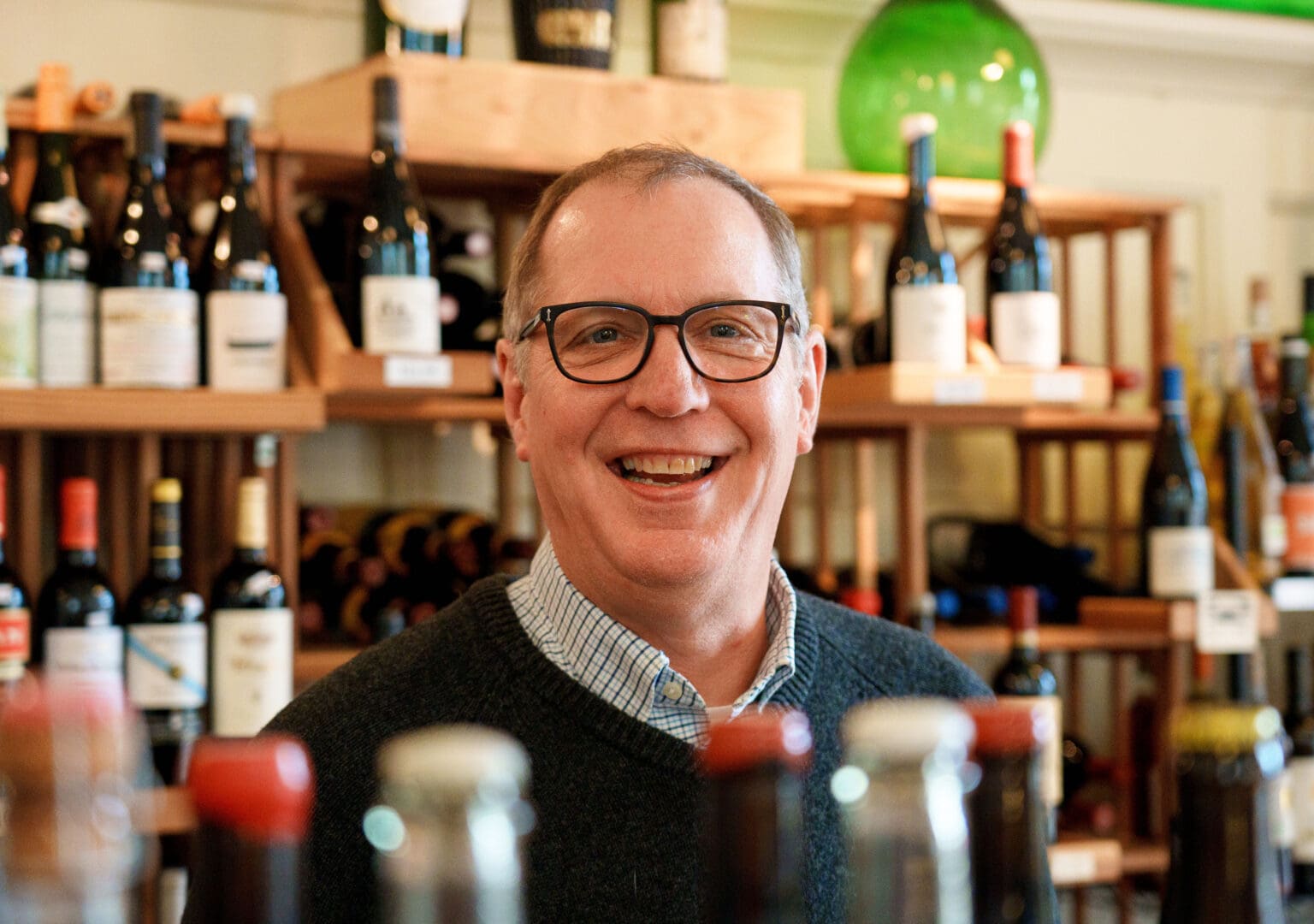Ted Seifert, owner of Seifert & Jones Wine Merchants smiles for the camera behind bottles of wine.