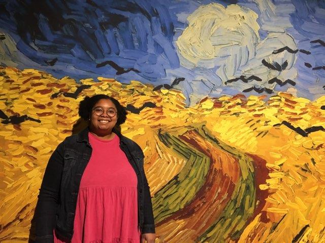 Artist Kayla Williams in front of a large painting, dressed in a bright pink dress and black jacket.