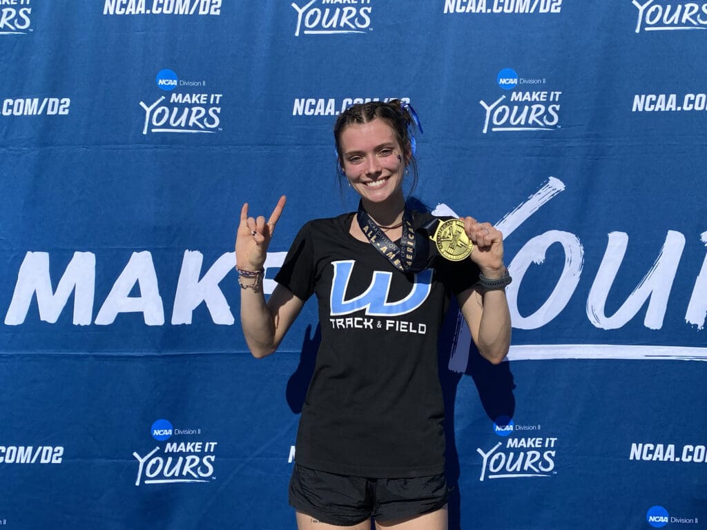 Western Washington University sophomore Ashley Reeck poses with her All-American medal as she holds up her medal and a rock gesture in front of a blue background.