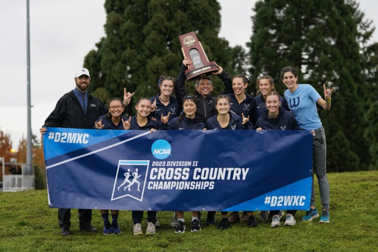 Western Washington University assistant coach and cross country lead T.J. Garlatz hoists the women's NCAA Division II West Regional championship trophy over the banner as the team smiles for the camera.