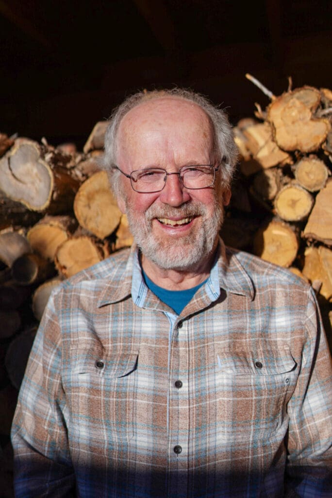 Author and institute insider John C. Miles smiles for the camera in front of logs stacked on top of each other.