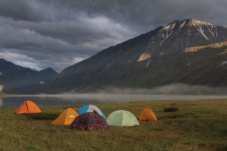 A group of colorful tents sit near a lakeshore in the Arctic National Wildlife Refuge as the sky is covered in dark gray clouds.