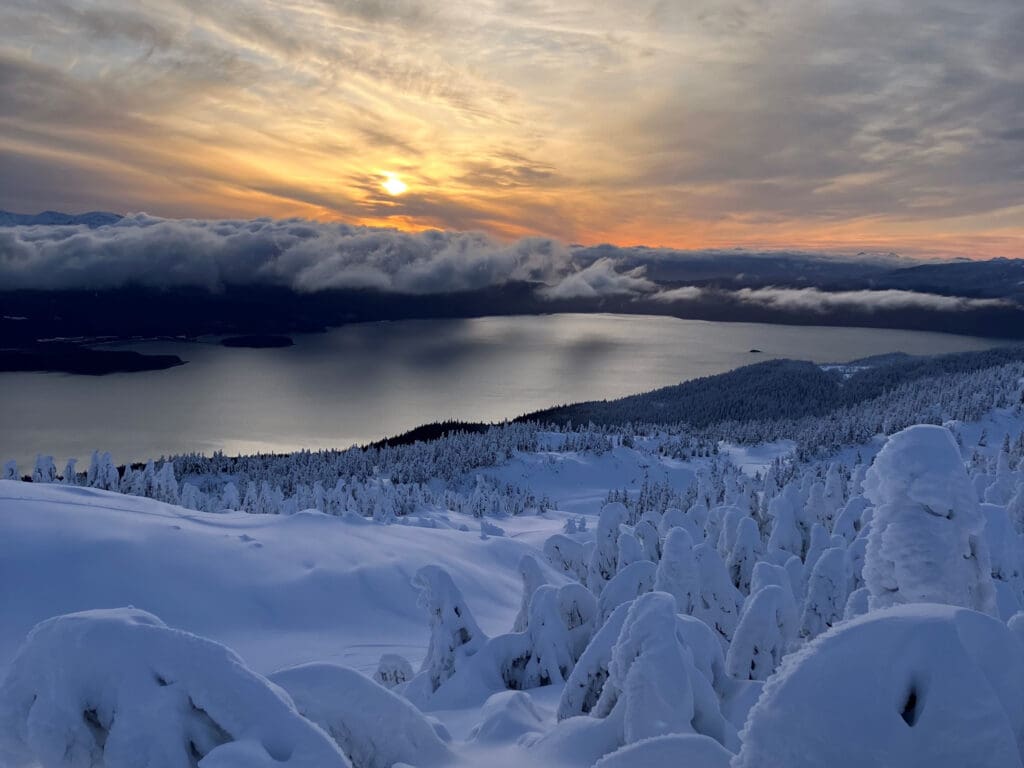 A scenic photo of mountains and trees covered in snow as clouds loom over a large body of water.