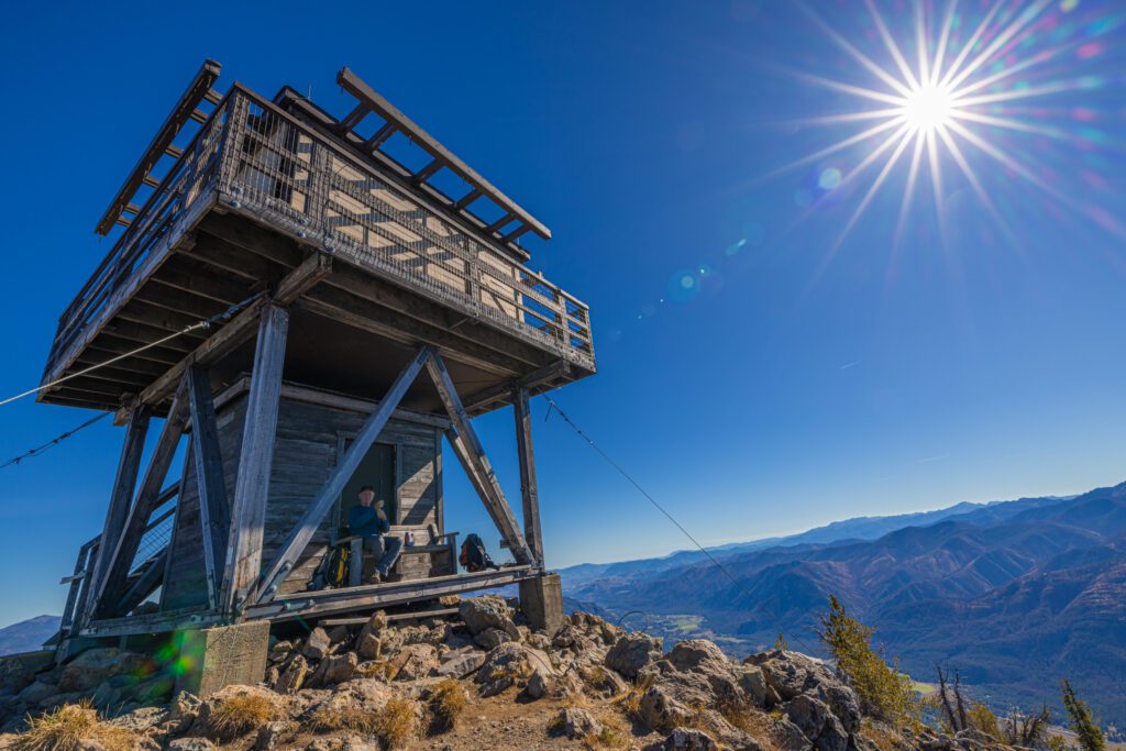 Elliott Almond breathes in 360-degree views at the Goat Peak fire lookout surrounded by rocks and the bright sun shining on the outpost.