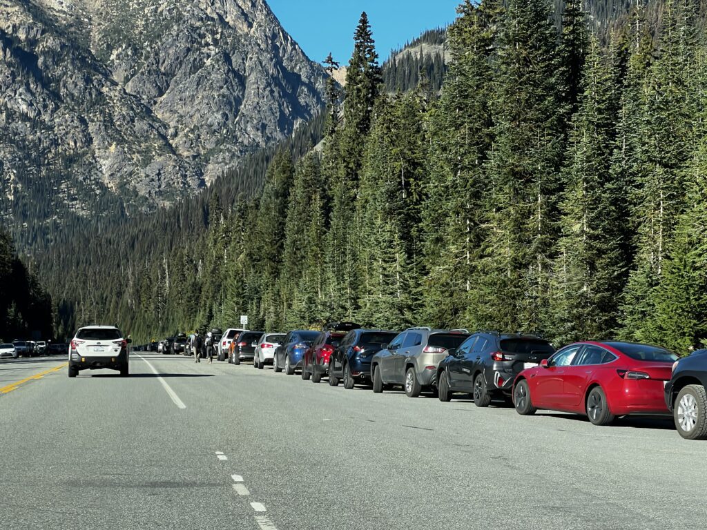 Parked cars line state Route 20 as people walk from their parking spot to the trailheads.